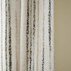 May include: Close-up of a textured fabric with vertical stripes. The fabric is mainly off-white with varying widths of black, grey, and beige stripes. The stripes have a slightly raised, textured appearance, creating a tactile visual effect. The fabric is likely used for home decor.