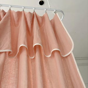 Puede incluir: Una cortina rosa pálido con un borde blanco y un borde superior festoneado. La cortina cuelga de una barra de metal blanco con clips plateados. La tela parece ligera y semitransparente, creando una estética delicada y aireada.