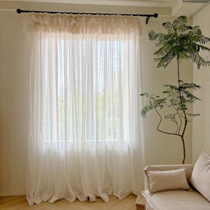 Puede incluir: Cortinas blancas transparentes cuelgan frente a una ventana, con una cenefa decorativa en la parte superior. Una barra de cortina negra sostiene las cortinas. Un pequeño taburete de madera está frente a un sofá beige con un cojín. Una planta en maceta está a la derecha.
