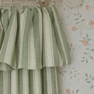 Puede incluir: Cortinas a rayas verdes y blancas colgadas de una barra crema con terminales decorativos. Las cortinas son de tela texturizada y están en capas. El fondo presenta un papel pintado floral.