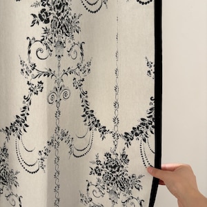 May include: Cream-colored fabric with a black floral pattern. The design features large floral bouquets, scrolling vines, and beaded accents. A black border frames the fabric's edge. The fabric is being held by a hand.