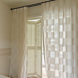 May include: Cream-coloured, sheer curtains with a chequered pattern hang from a black rod above a window with white shutters. The curtains are partially drawn, allowing light to filter through. The room has a neutral colour palette.