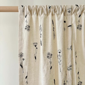 Op de afbeelding: Crèmekleurig gordijnpaneel met een herhalend patroon van zwarte bloemenlijntekeningen. Het gordijn is aan de bovenkant geplooid en hangt aan een houten roede. De bloemenontwerpen omvatten verschillende bloemen- en plantillustraties.