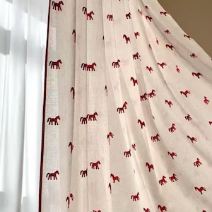 May include: White curtain with a repeating pattern of red horse silhouettes. The curtain has a red border and is hanging in front of a window. The fabric is slightly translucent.
