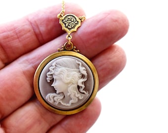 May include: A round pendant necklace featuring a white and grey carved cameo profile of a person with flowing hair, set in a gold-toned metal frame. The pendant is attached to a decorative gold-toned bail with an ornate scrollwork design.