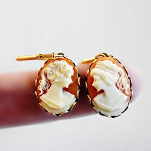 May include: A pair of oval-shaped earrings featuring a carved white profile of a person on a light orange background. Each cameo is encased in a thin gold-toned metal frame with a lever-back closure mechanism attached to the top of the setting.