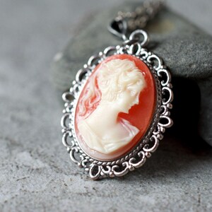 May include: A silver pendant necklace with a cameo featuring a woman's profile in white against a coral background. The cameo is surrounded by a decorative silver frame.