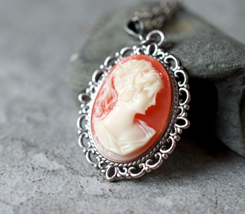 May include: A silver pendant necklace with a cameo featuring a woman's profile in white against a coral background. The cameo is surrounded by a decorative silver frame.