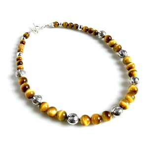 May include: A beaded necklace featuring round, golden-brown tiger's eye gemstones interspersed with polished silver-toned metal beads. The necklace is secured with a silver-toned toggle clasp, creating a circular arrangement of alternating stone and metal.