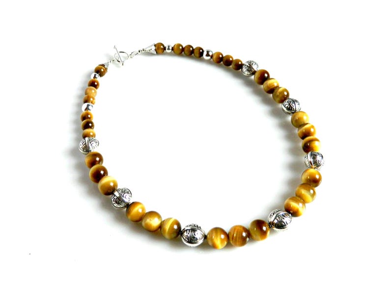 May include: A beaded necklace featuring round, golden-brown tiger's eye gemstones interspersed with polished silver-toned metal beads. The necklace is secured with a silver-toned toggle clasp, creating a circular arrangement of alternating stone and metal.