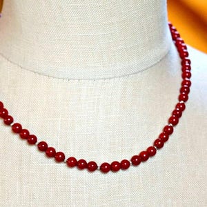May include: A red coral bead necklace on a white mannequin. The necklace is made of small, round beads that are strung together on a silver chain.