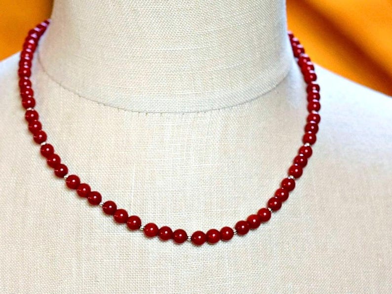 May include: A red coral bead necklace on a white mannequin. The necklace is made of small, round beads that are strung together on a silver chain.