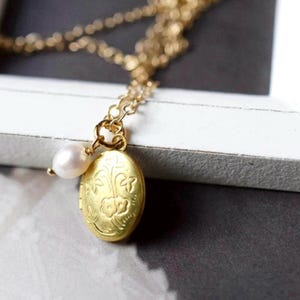 May include: A gold-colored necklace with a small, oval locket pendant and a single pearl. The locket features a floral design and hangs from a delicate chain. The necklace is displayed on a white surface.