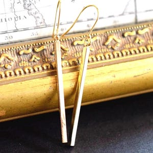 May include: A pair of gold-colored, linear drop earrings. Each earring features a long, rectangular bar suspended from a curved ear wire. The earrings are displayed against a backdrop of a vintage map and a decorative gold frame.