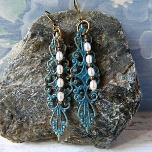 May include: Teal blue filigree earrings with white pearl beads hanging from the bottom. The earrings are on a gray rock.