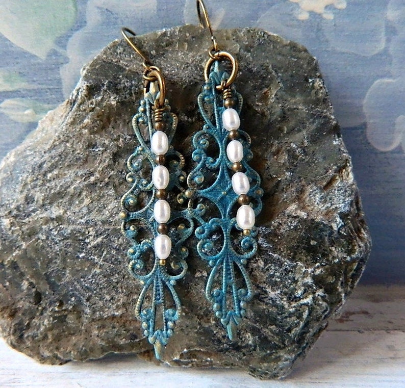 May include: Teal blue filigree earrings with white pearl beads hanging from the bottom. The earrings are on a gray rock.