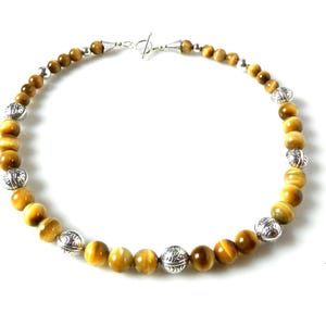 May include: A beaded necklace featuring round, golden-brown tiger's eye gemstones interspersed with decorative, etched silver-toned metal beads. The necklace is secured with a silver-toned toggle clasp at the top, creating a circular arrangement.