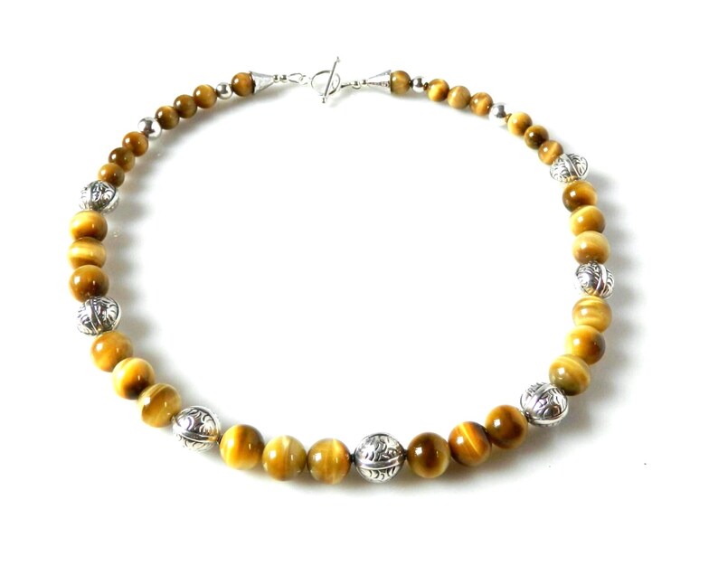 May include: A beaded necklace featuring round, golden-brown tiger's eye gemstones interspersed with decorative, etched silver-toned metal beads. The necklace is secured with a silver-toned toggle clasp at the top, creating a circular arrangement.