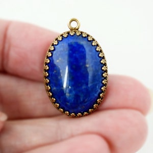 May include: An oval pendant featuring a deep blue lapis lazuli stone with gold flecks, encased in a decorative antique brass metal setting with a scalloped edge and a small circular jump ring at the top for attachment to a chain or cord.