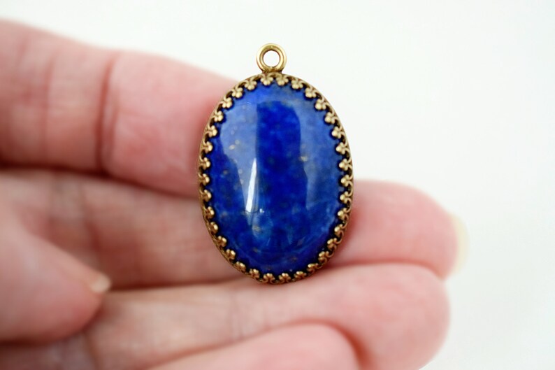 May include: An oval pendant featuring a deep blue lapis lazuli stone with gold flecks, encased in a decorative antique brass metal setting with a scalloped edge and a small circular jump ring at the top for attachment to a chain or cord.