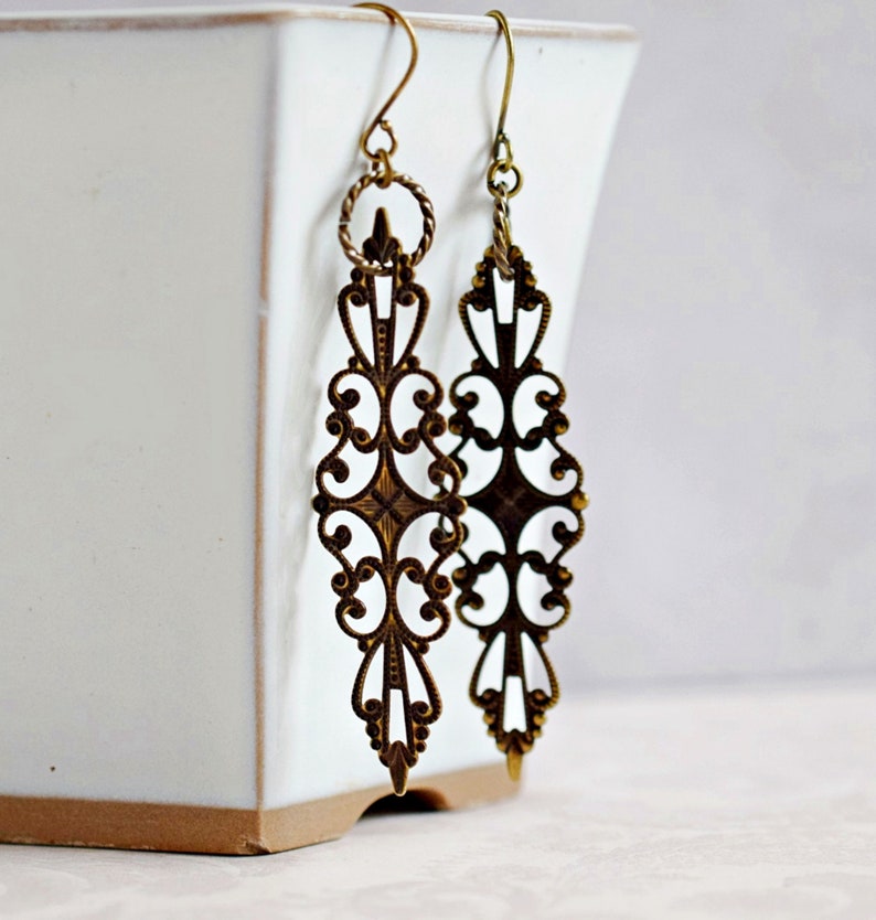 May include: A pair of long, gold-tone earrings with a delicate, intricate filigree design. The earrings are made of metal and have a dangling, drop shape.