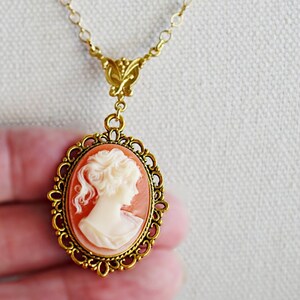 May include: A gold chain necklace with a cameo pendant. The cameo is a portrait of a woman with a white background and a peach-colored frame. The frame is decorated with a gold filigree design.
