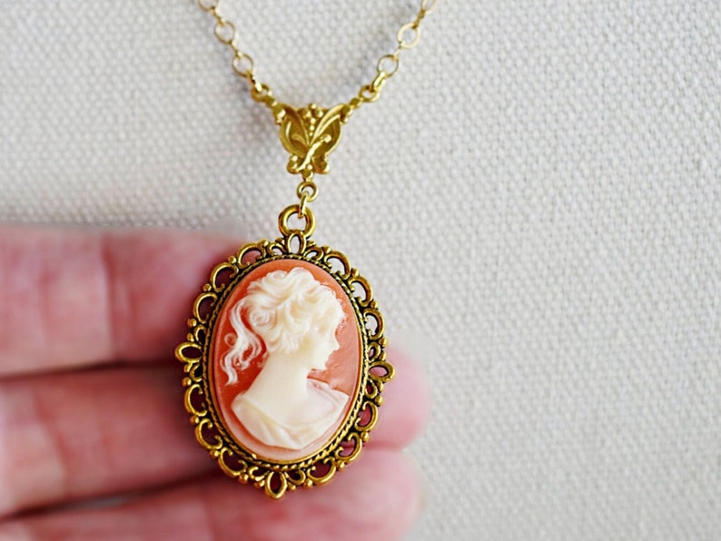 May include: A gold chain necklace with a cameo pendant. The cameo is a portrait of a woman with a white background and a peach-colored frame. The frame is decorated with a gold filigree design.