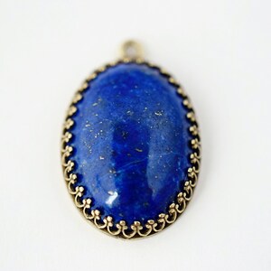 May include: An oval pendant featuring a deep blue lapis lazuli stone with gold-colored flecks, set within a decorative antique brass-toned metal frame with a scalloped edge and a small circular bail at the top for attachment to a chain.
