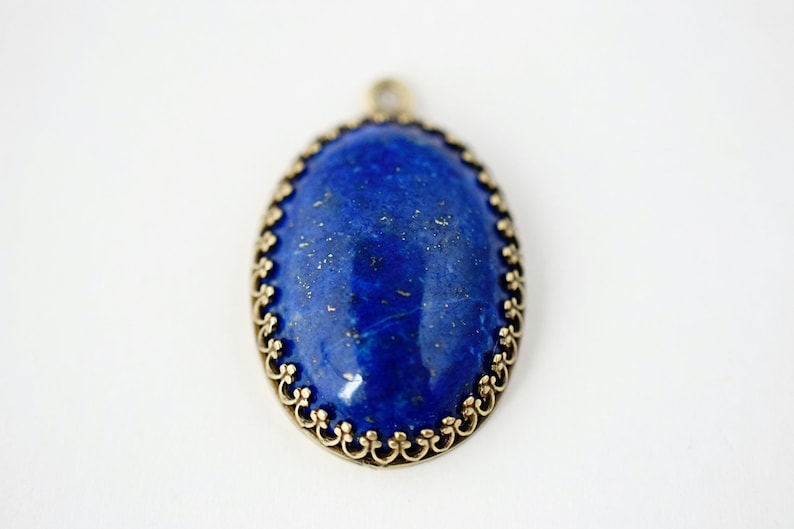 May include: An oval pendant featuring a deep blue lapis lazuli stone with gold-colored flecks, set within a decorative antique brass-toned metal frame with a scalloped edge and a small circular bail at the top for attachment to a chain.