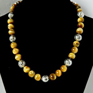 May include: A beaded necklace featuring alternating round golden-brown tiger eye gemstone beads and smaller, spherical silver-toned metal beads with an intricate engraved pattern, arranged in a repeating sequence along the entire length of the strand.
