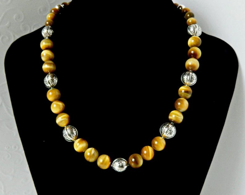 May include: A beaded necklace featuring alternating round golden-brown tiger eye gemstone beads and smaller, spherical silver-toned metal beads with an intricate engraved pattern, arranged in a repeating sequence along the entire length of the strand.