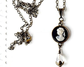 Puede incluir: Un collar de cadena de bronce con un colgante de camafeo negro que presenta una silueta blanca de una mujer. Una lágrima de cristal transparente cuelga de la parte inferior del colgante.