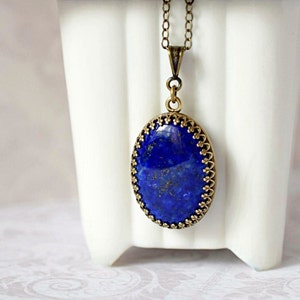 May include: An oval lapis lazuli pendant featuring deep blue stone with gold-colored flecks, set in an ornate, antique bronze-toned metal frame with a scalloped edge. The pendant hangs from a matching bronze-toned metal chain link necklace.