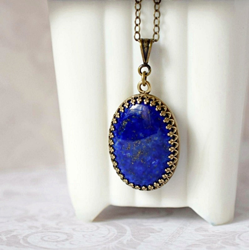 May include: An oval lapis lazuli pendant featuring deep blue stone with gold-colored flecks, set in an ornate, antique bronze-toned metal frame with a scalloped edge. The pendant hangs from a matching bronze-toned metal chain link necklace.