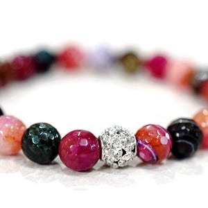 May include: A beaded bracelet featuring a variety of colorful, round beads. The beads are in shades of pink, red, green, and black, with a silver-colored, rhinestone-studded bead. The bracelet is on a white background.