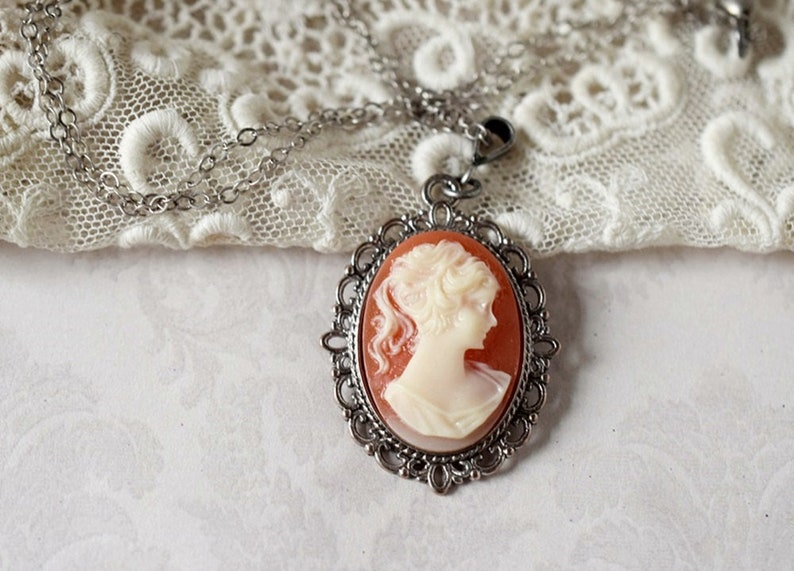 May include: A silver chain necklace with a cameo pendant. The cameo features a white profile of a woman with a detailed hairstyle, set against a peach-colored background. The cameo is surrounded by a silver filigree frame.