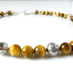 May include: A necklace featuring round, golden-brown tiger's eye beads alternating with polished, etched silver-toned metal beads. The strand is arranged in a circular shape and includes a silver-toned metal clasp mechanism at the back.