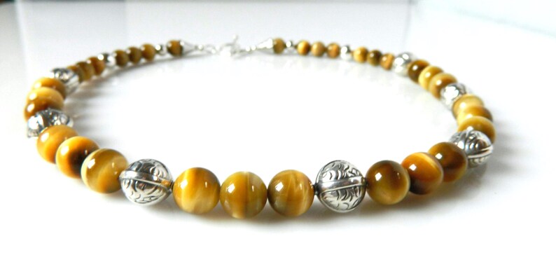 May include: A necklace featuring round, golden-brown tiger's eye beads alternating with polished, etched silver-toned metal beads. The strand is arranged in a circular shape and includes a silver-toned metal clasp mechanism at the back.