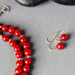 May include: A red beaded necklace and matching earrings. The necklace is made of round, red beads and has a silver clasp. The earrings are silver with red beads hanging from them.