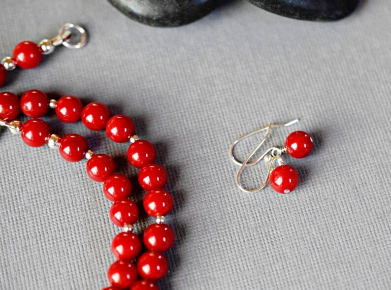 May include: A red beaded necklace and matching earrings. The necklace is made of round, red beads and has a silver clasp. The earrings are silver with red beads hanging from them.