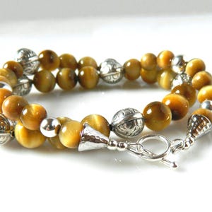 May include: A double-strand bracelet featuring polished golden-brown tiger's eye beads interspersed with round, etched silver-tone metal beads. The piece is finished with a silver-tone toggle clasp and conical end caps, creating a layered look.