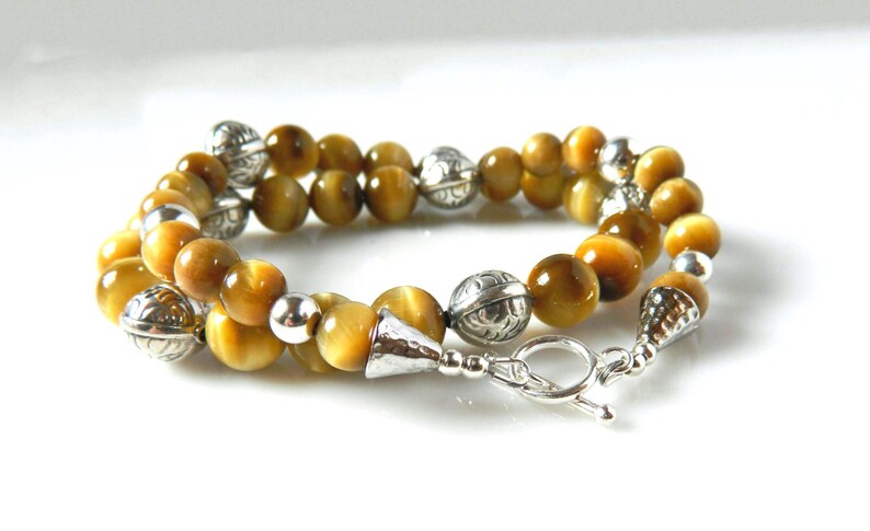 May include: A double-strand bracelet featuring polished golden-brown tiger's eye beads interspersed with round, etched silver-tone metal beads. The piece is finished with a silver-tone toggle clasp and conical end caps, creating a layered look.