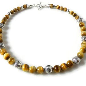 May include: A beaded necklace featuring round, golden-brown tiger's eye gemstones alternating with spherical, silver-toned metal beads that have an intricate engraved pattern. The necklace is secured with a silver-toned toggle clasp closure.
