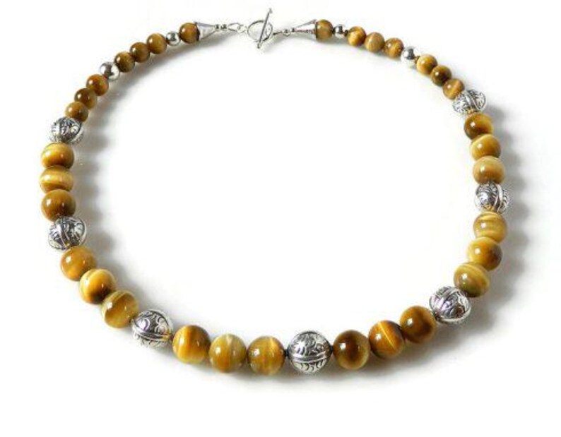 May include: A beaded necklace featuring round, golden-brown tiger's eye gemstones alternating with spherical, silver-toned metal beads that have an intricate engraved pattern. The necklace is secured with a silver-toned toggle clasp closure.