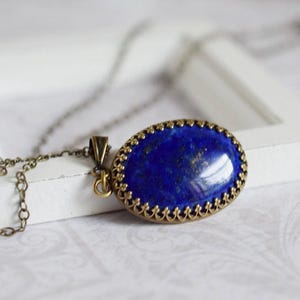 May include: An oval-shaped lapis lazuli pendant featuring a deep blue stone with subtle gold-colored flecks, set in an ornate, antique-style bronze metal frame with a scalloped edge, attached to a matching thin bronze metal link chain necklace.