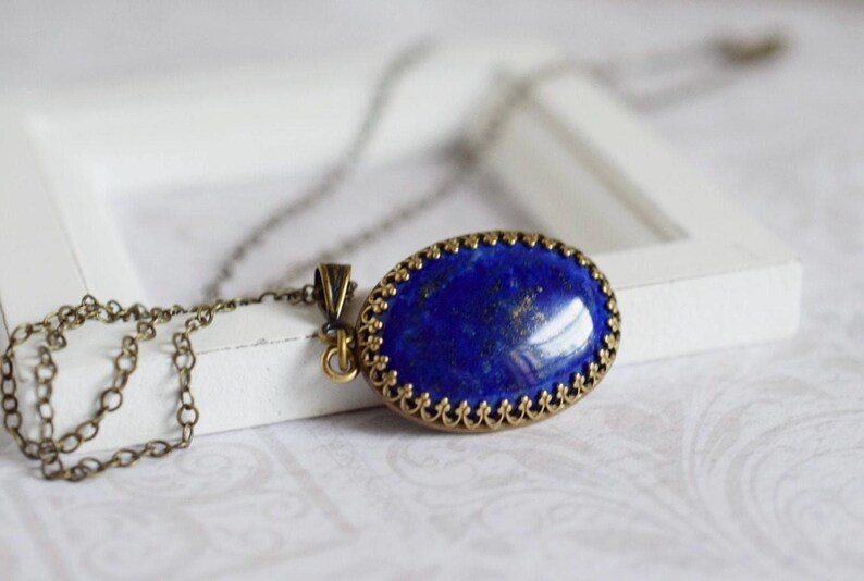 May include: An oval-shaped lapis lazuli pendant featuring a deep blue stone with subtle gold-colored flecks, set in an ornate, antique-style bronze metal frame with a scalloped edge, attached to a matching thin bronze metal link chain necklace.