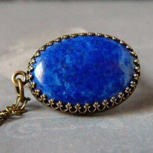 May include: An oval pendant featuring a deep blue lapis lazuli stone with natural flecks, set in a decorative bronze-toned metal frame with a scalloped edge. The pendant is attached to a matching bronze-toned metal chain link necklace.