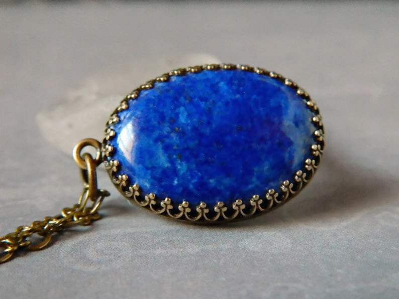 May include: An oval pendant featuring a deep blue lapis lazuli stone with natural flecks, set in a decorative bronze-toned metal frame with a scalloped edge. The pendant is attached to a matching bronze-toned metal chain link necklace.
