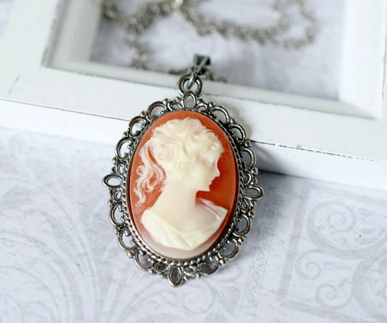 May include: A silver chain necklace with a cameo pendant. The cameo is an oval shape with a white profile of a woman against a peach background.