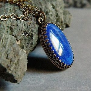 May include: An oval-shaped blue lapis lazuli gemstone pendant set in a decorative bronze-toned metal bezel. The pendant hangs from a matching bronze-toned metal chain, featuring a smooth, polished surface that reflects light across its deep blue hue.